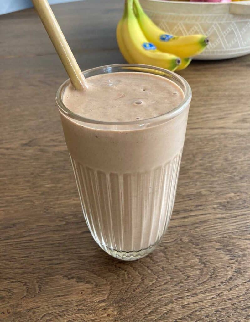 Peanut butter avocado smoothie in clear cup with a brown straw. A bunch of bananas are in the background. Smoothie sits on a wood table.