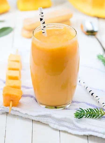 Orange smoothie in clear glass on a white towel on a white picnic table surrounded by papaya cubes on a stick.