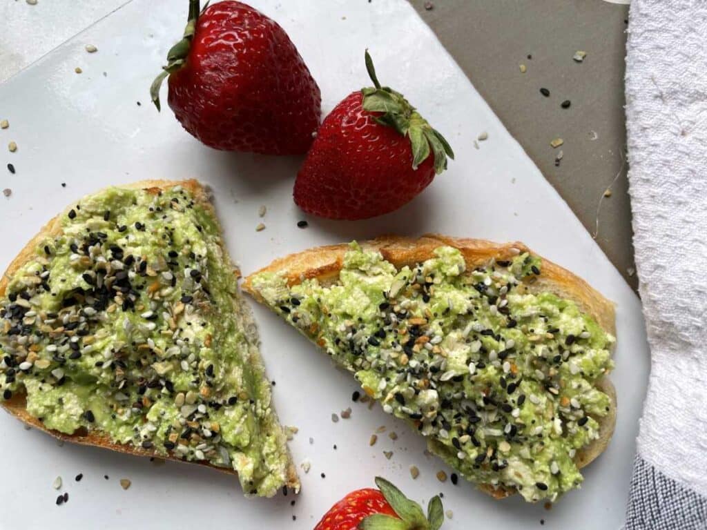 A slice of tofu toast on white cutting board surrounded by 3 strawberries. White toast cut in half, with avocado, tofu and bagel spice on top.