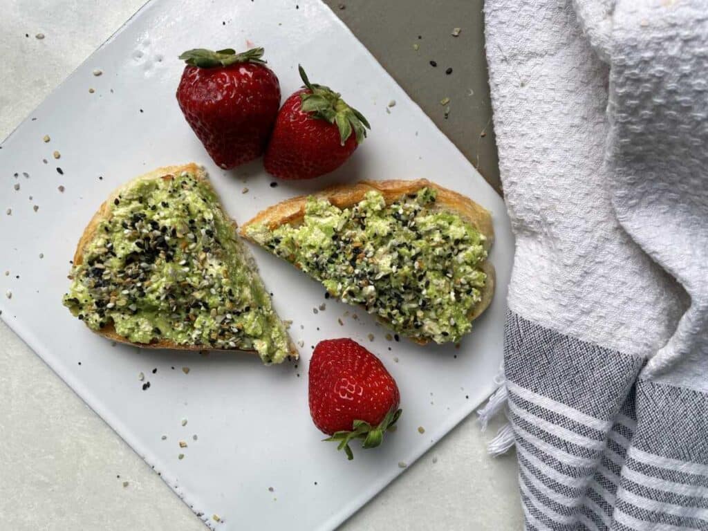 A slice of tofu toast on white cutting board surrounded by 3 strawberries. White toast cut in half, with avocado, tofu and bagel spice on top.
