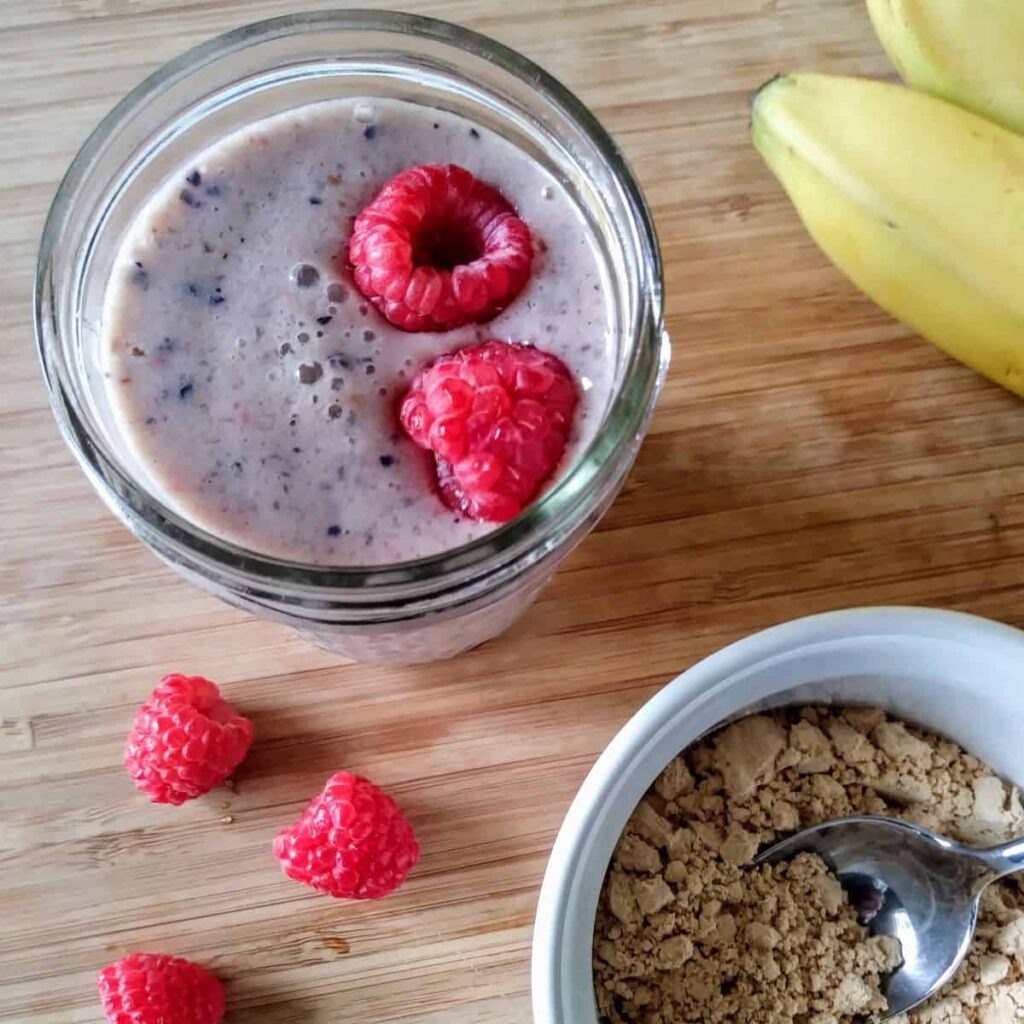 DASH smoothie that is purple in color with two raspberries floating on top and three surrounding it with banana and flaxseed in the corner of a wood table.