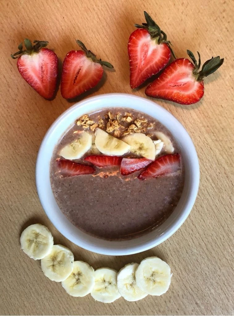 DASH smoothie bowl that is brown toped with sliced strawberries, bananas and walnuts surrounded by sliced strawberries and bananas.