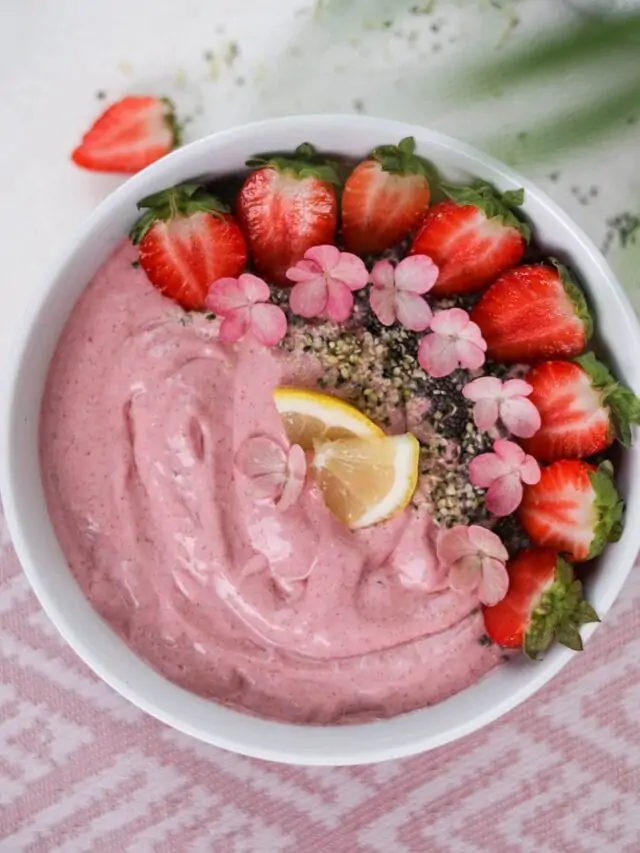 Pink smoothie bowl topped with sliced strawberries, pink flowers, herbs and sliced lemons. 