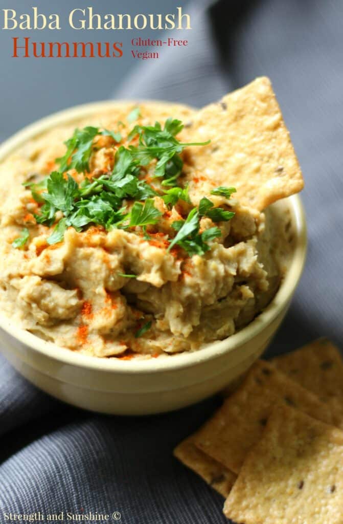 Baba ghanoush hummus in a white bowl with green herbs and a cracker sticking out.