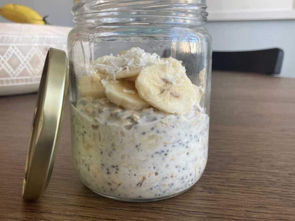 Banana cream overnight oats in a glass jar, topped wiht sliced bananas and coconut flakes. Lid of glass jar is leaning against the jar on a wood table with a kitchen chair in the background.