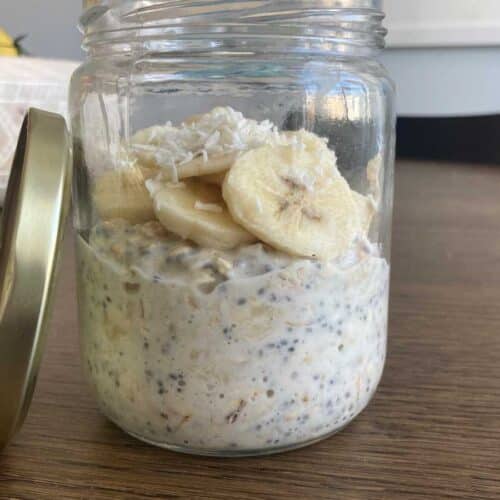Banana cream overnight oats in a glass jar, topped wiht sliced bananas and coconut flakes. Lid of glass jar is leaning against the jar on a wood table with a kitchen chair in the background.