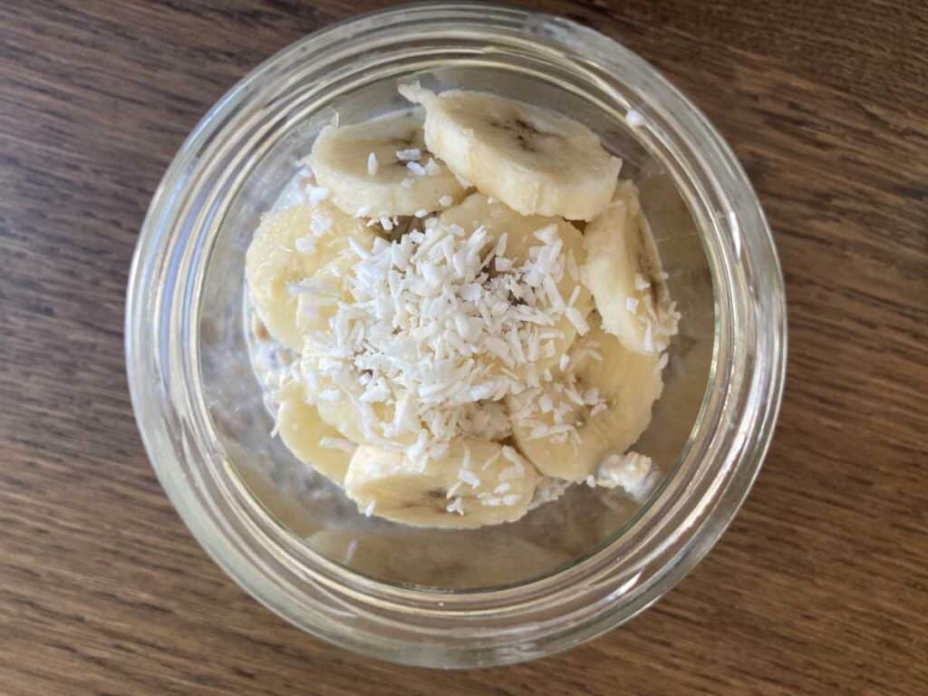 Banana cream overnight oats in a glass jar with a view from above showing the toppings of sliced bananas and shredded coconut.