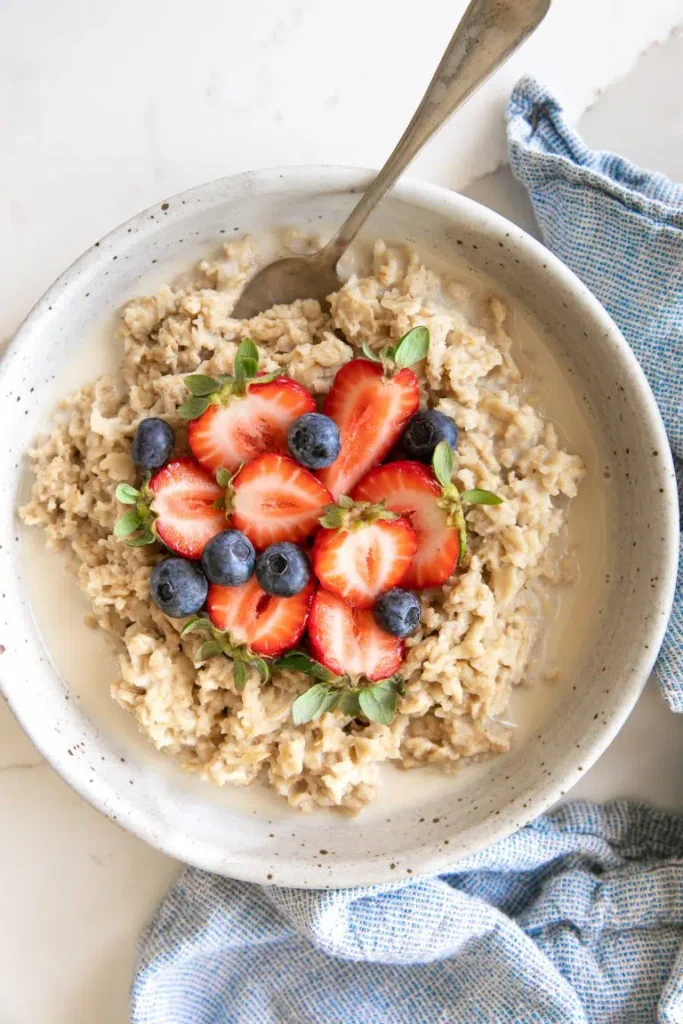 A bowl of oatmeal topped with strawberries, blueberries, and a spoon, beside a blue tea towel. 