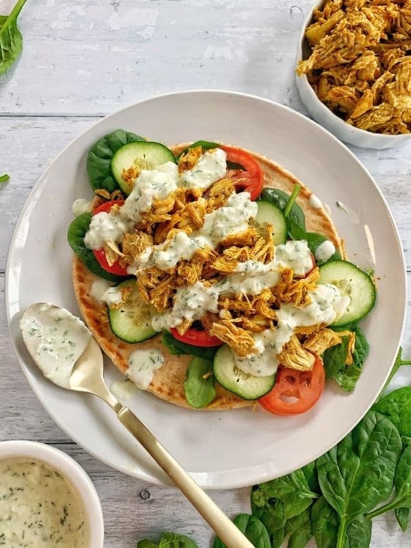 Low sodium instant pot chicken shawarma wrap on a white pita, drizzled with tzatziki dressing. Displayed on a white plate with a used spoon and shredded chicken, tzatziki dip and basil are in the background.