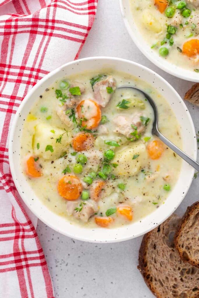 A large bowl of low sodium instant pot of creamy chicken stew with a spoon, beside a red and white tea towel and brown bread.