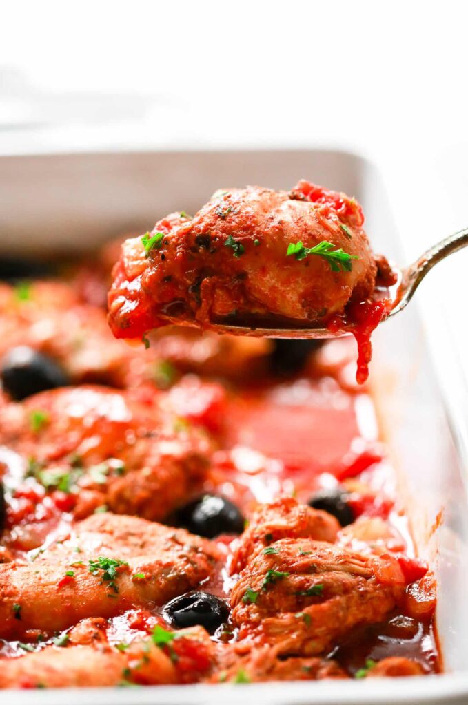 Low sodium instant pot chicken in a casserole dish with olives, tomato and green herbs.  One chicken thigh is elevated on a serving spoon.