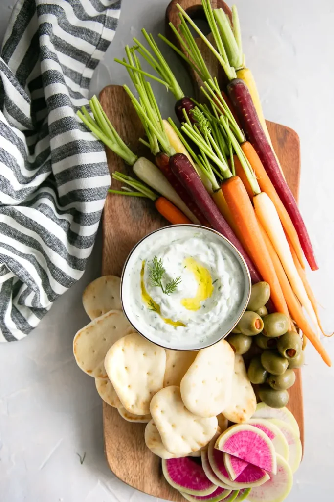 Tzatziki sauce in a white bowl on a wood plank with olives, sliced radishes, crackers and whole carrots with the green stems. 