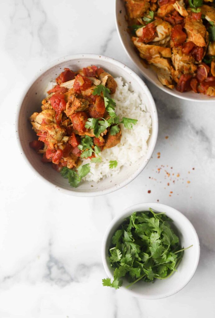 Low sodium instant pot chicken and tomato on a bed of white rice in a small white bowl, garnished with green herbs, beside a large white bowl of chicken and tomatoes and a smaller white bowl of green herbs.