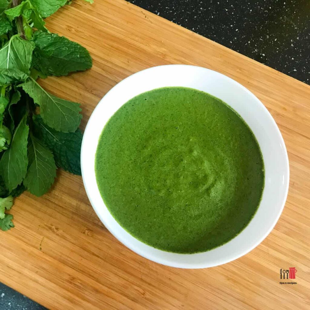 Green chutney in a white bowl on a wood cutting board beside green herbs.