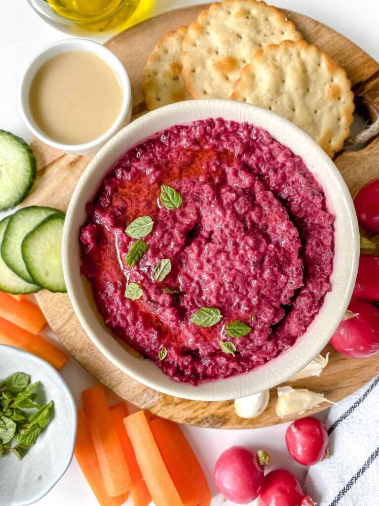 Low sodium pink hummus in a white bowl topped with green herbs on a snack board filled with cucumber slices, carrot sticks, radishes and crackers.