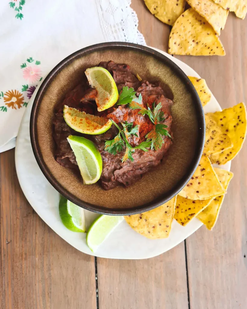 Black bean low sodium dip in a grey bowl topped with slices of lime, herbs and chipotle pepper.  The bowl is surrounded by corn chips and a white napkin. 