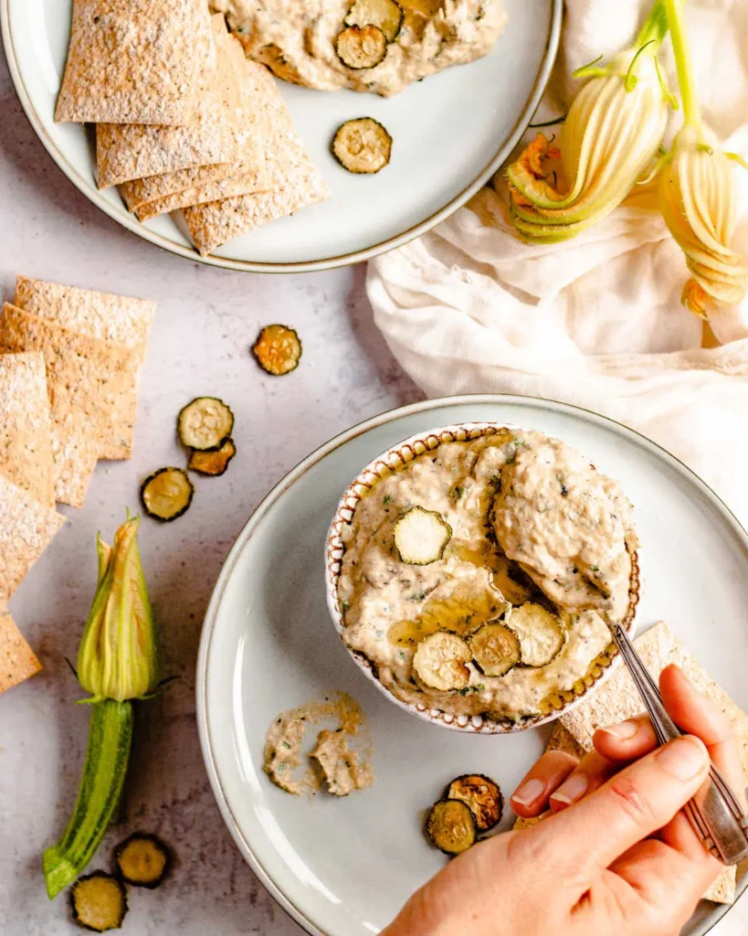 Babaganoush in a patterned bowel with sliced cucumber on top, with a just dipped spoon being held by a hand. This Babaganoush is on a white plate surrounded by crackers, flowers and sliced cucumbers.