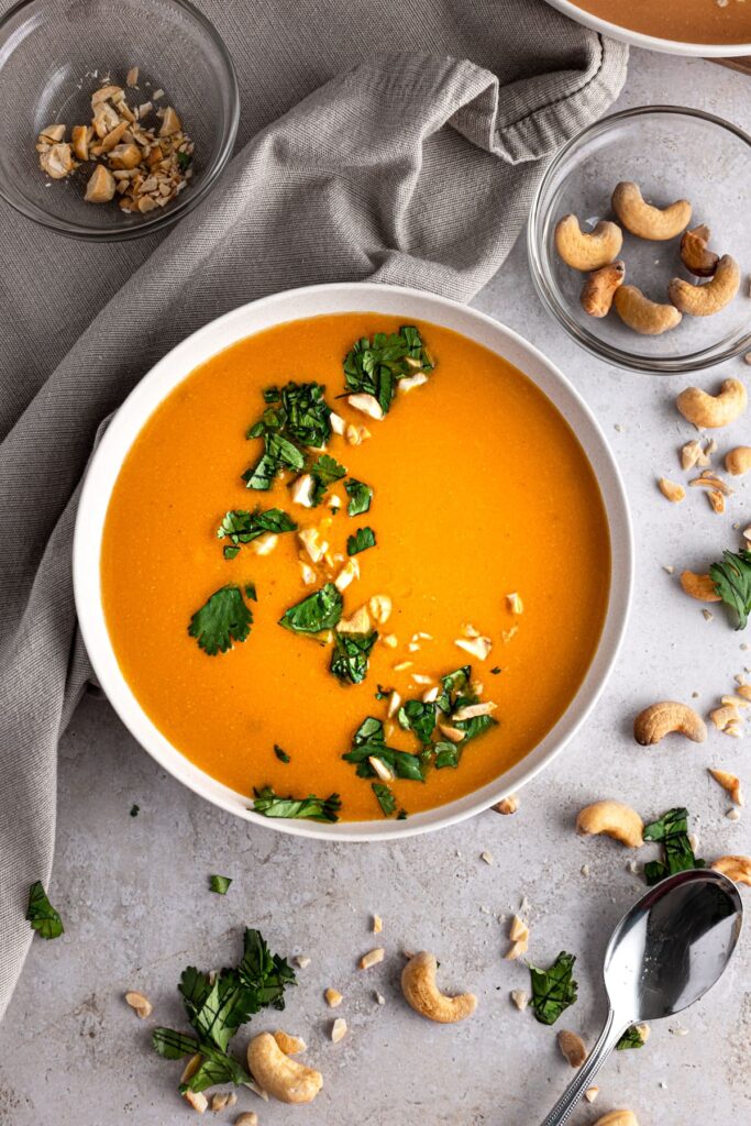 Bring orange soup in a white bowl topped with broken cashews and green herbs beside a grey towel.