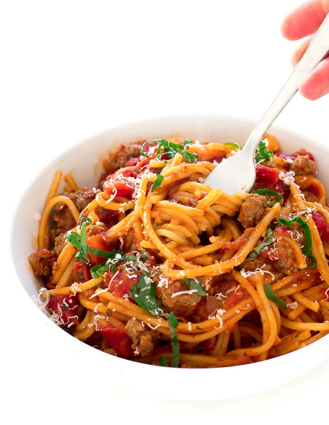 White bowl of spaghetti ground meat, tomatoes and fresh herbs and a loaded fork. 