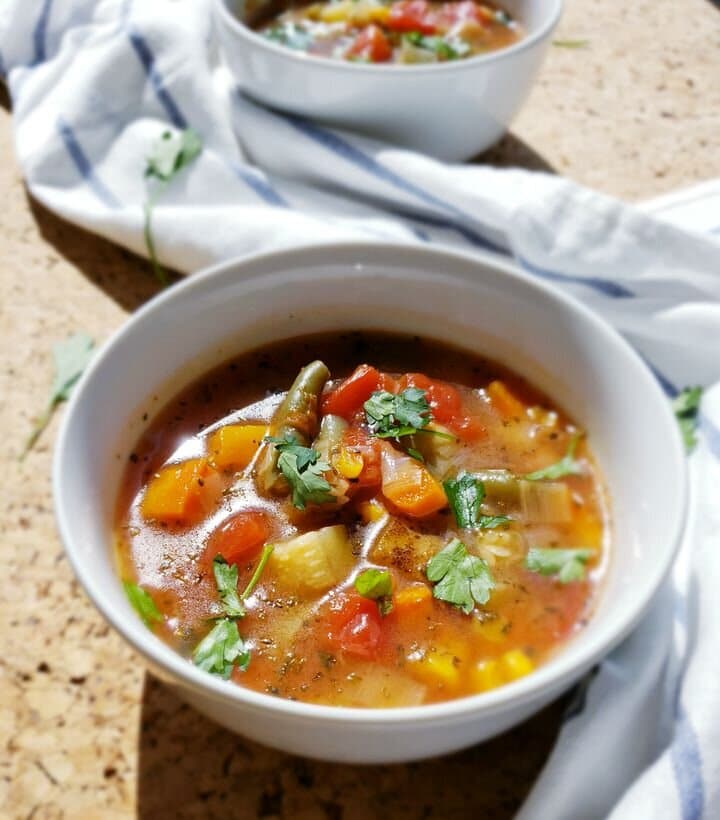 2 bowls of vegetable soup topped with fresh herbs in white bowls, surrounded by white tea towel.