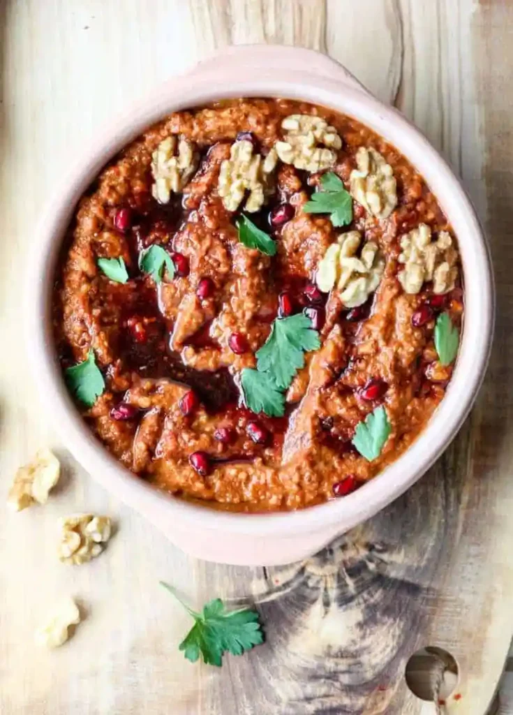 Red pepper low sodium dip in a white bowl topped with pomegranate, walnuts and green herbs.