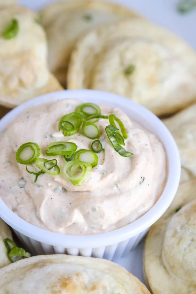 White dip in a white container topped with sliced green onion surrounded by pita.  
