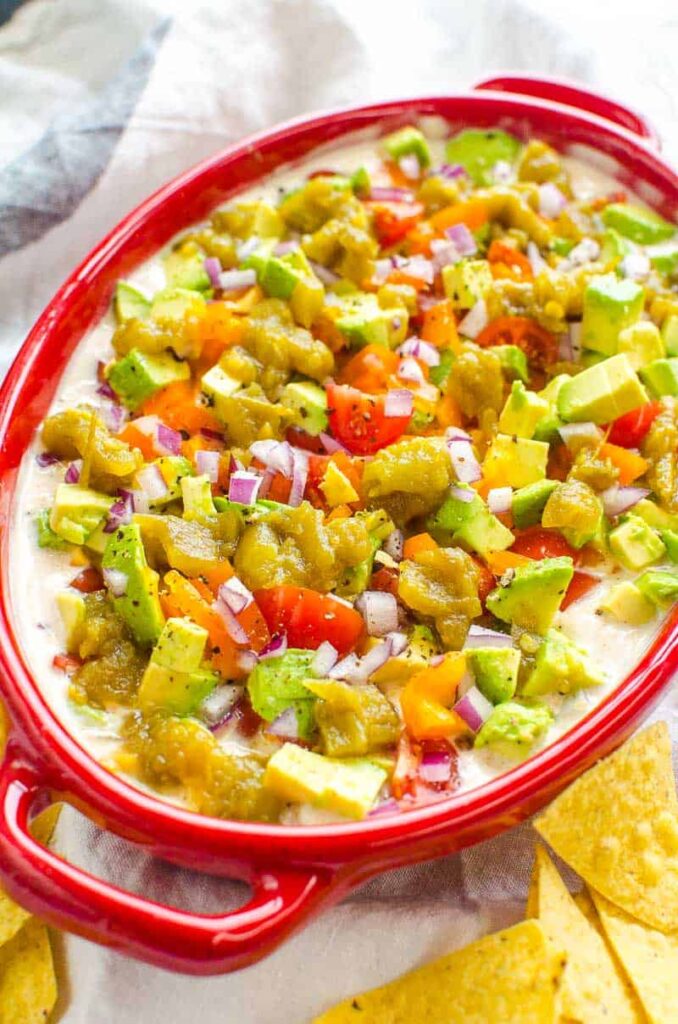 Low sodium taco dip with lots of toppings in a red bowl over a blue and white napkin that is sprinkled with corn chips.