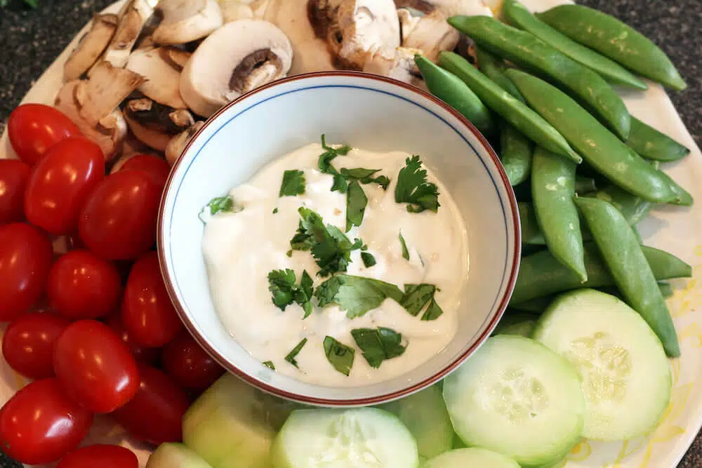 Low sodium dip for veggies in a white bowl topped with cilantro surrounded by vegetables including cherry tomatoes, sliced mushrooms, peas and cucumbers. 