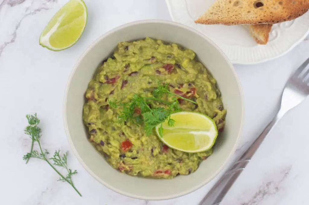 Low sodium appetizer of avocado topped with lime and fresh dill in a white bowl on a white marble counter beside a white plate and a fork. 