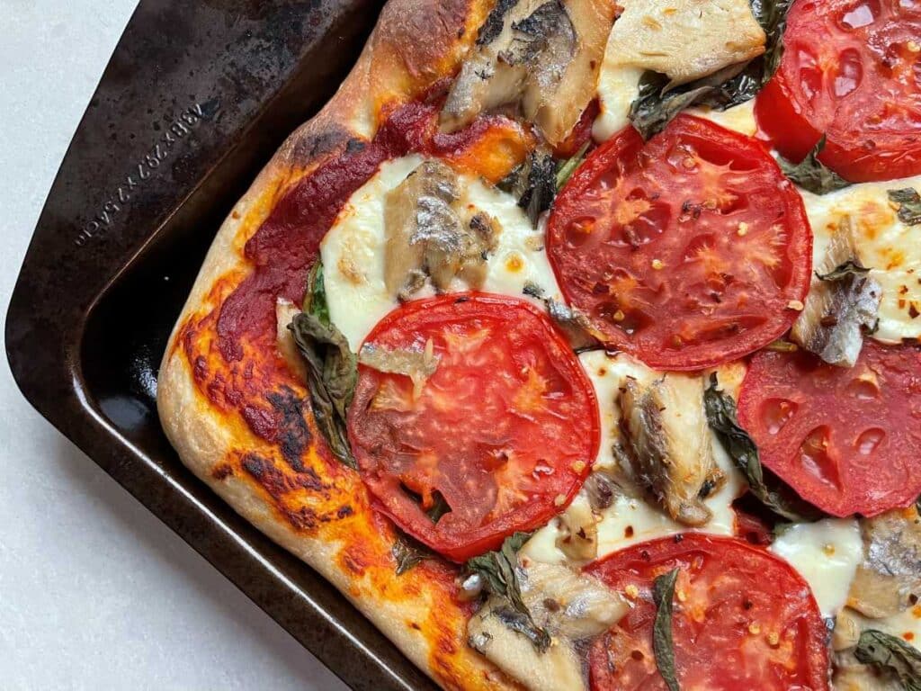 A corned of the cooked sardine pizza on baking sheet, with browned pizza crust, melted mozzarella cheese, crispy tomato slices and large chunks of sardines, on a white counter top.