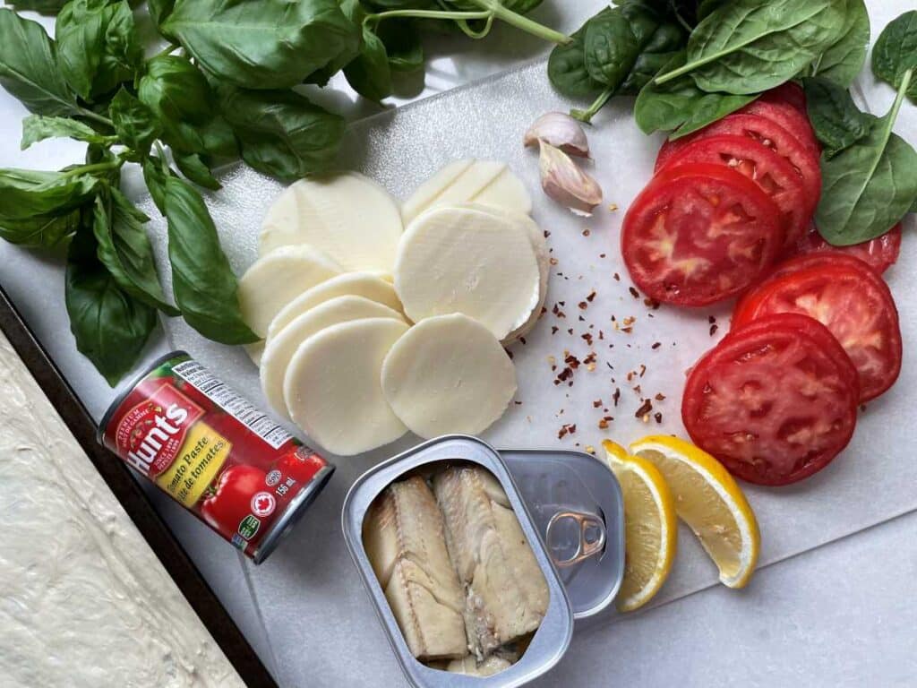 Ingredients for sardine pizza laid on white countertop include pizza dough spread in baking pan, tomato paste in can, sardines in tin with lid removed, 2 lemon wedges, tomato slices, baby spinach, whole fresh basil, garlic cloves, sliced mozzarella and sprinkle of red pepper flakes.