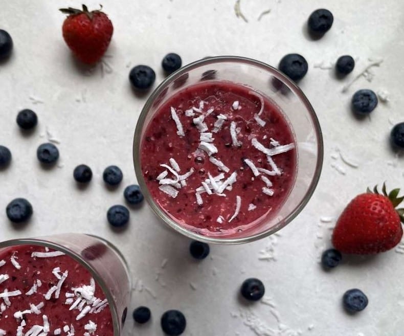 Two triple berry oat tropical smoothie in clear glasses surrounded by strawberries and blueberries on a white countertop.