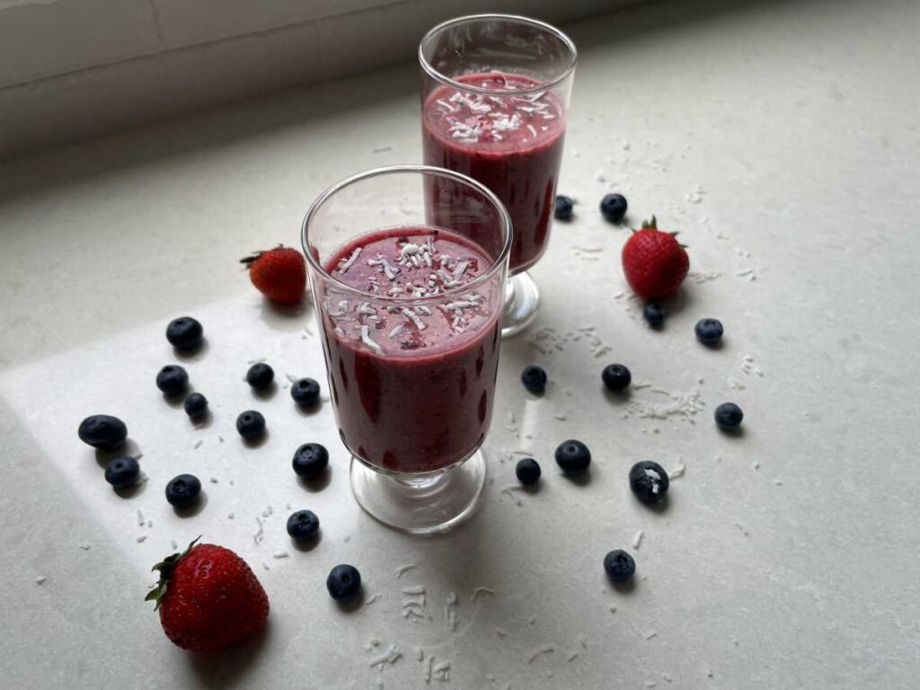 Two triple berry oat tropical smoothie in clear glasses surrounded by strawberries and blueberries on a white countertop.
