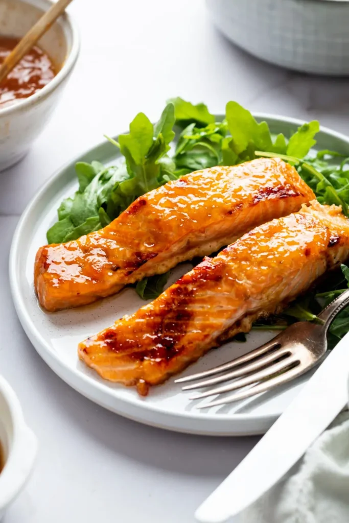 Two portions of salmon topped with orange marinade on a bed of green lettuce beside a fork on a white plate.