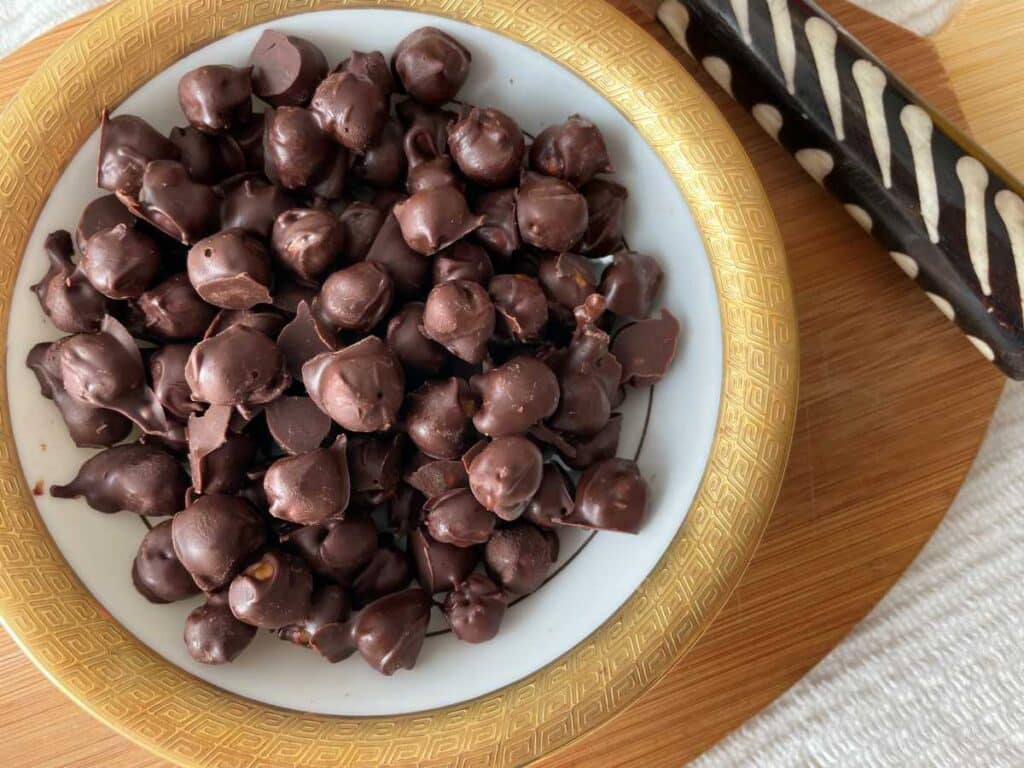 Chocolate covered chickpeas in a gold rimmed bowl over a wood cutting board beside a white and brown serving utensil.