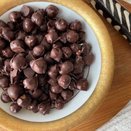 Chocolate covered chickpeas in a gold rimmed bowl over a wood cutting board beside a white and brown serving utensil.