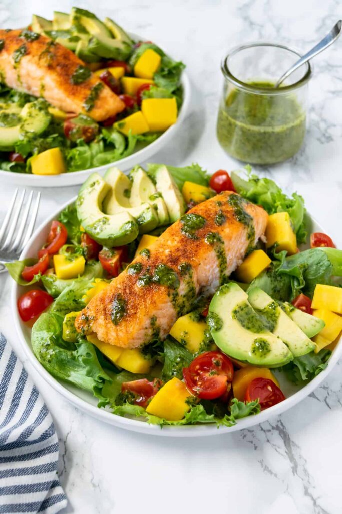 Two large white bowls of green salad topped with avocado, cherry tomatoes, and salmon drizzled with a green sauce.