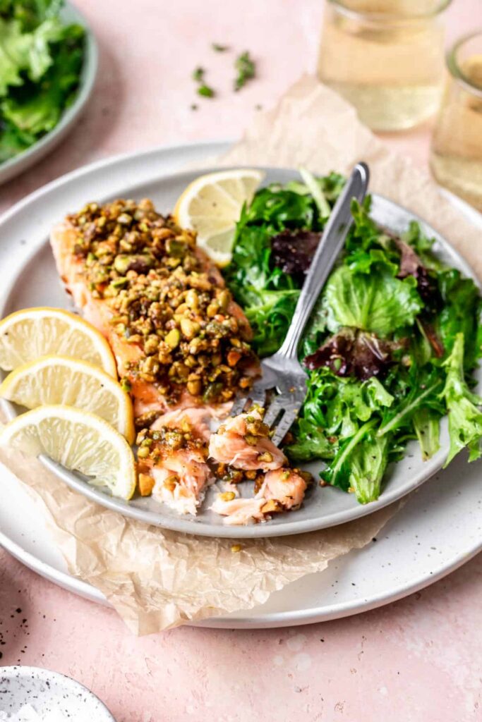 Pistachio Crusted Salmon fillet cut into wiht a fork on a white plate beside a green salad and lemon slices.