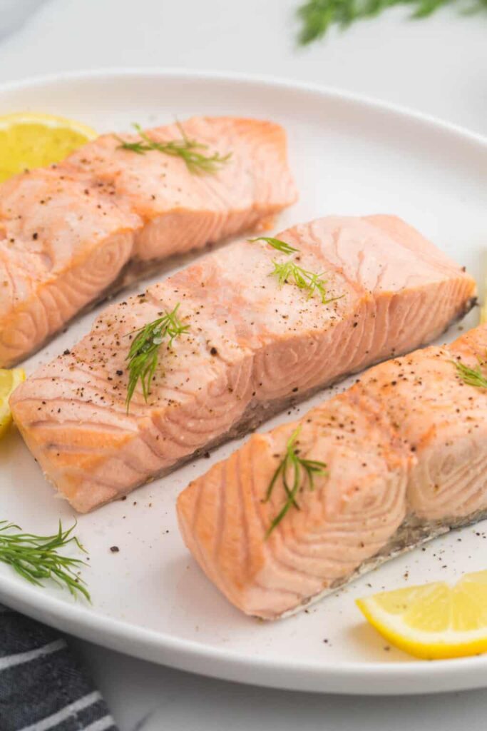 Thick salmon fillets with fresh dill and pepper on a white plate beside sliced lemon.