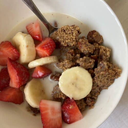 Chunky steel cut oat granola topped with sliced banana, strawberries and milk. In a white bowl with a spoon.