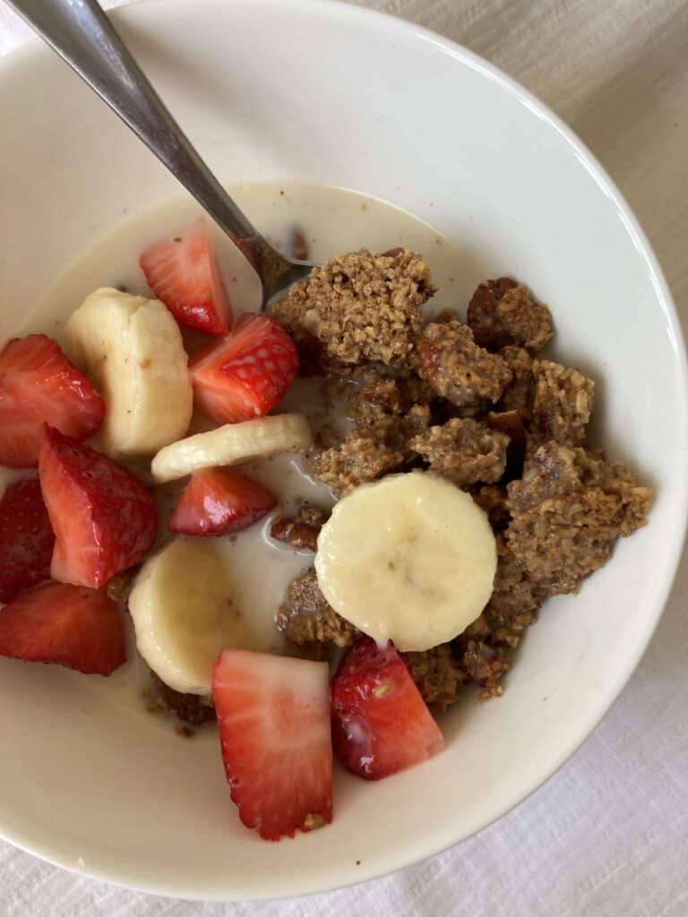 Chunky steel cut oat granola topped with sliced banana, strawberries and milk. In a white bowl with a spoon.