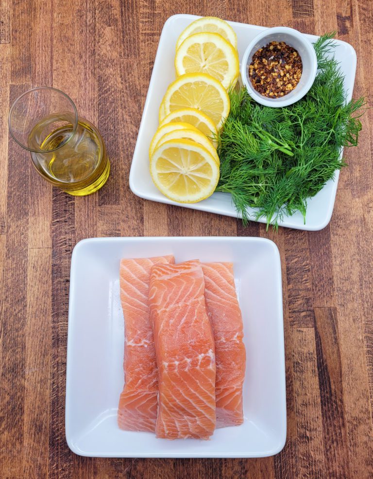Raw salmon on white plate, beside fresh lemon, fresh dill and chili peppers on another white plate, and a clear jar of olive oil.