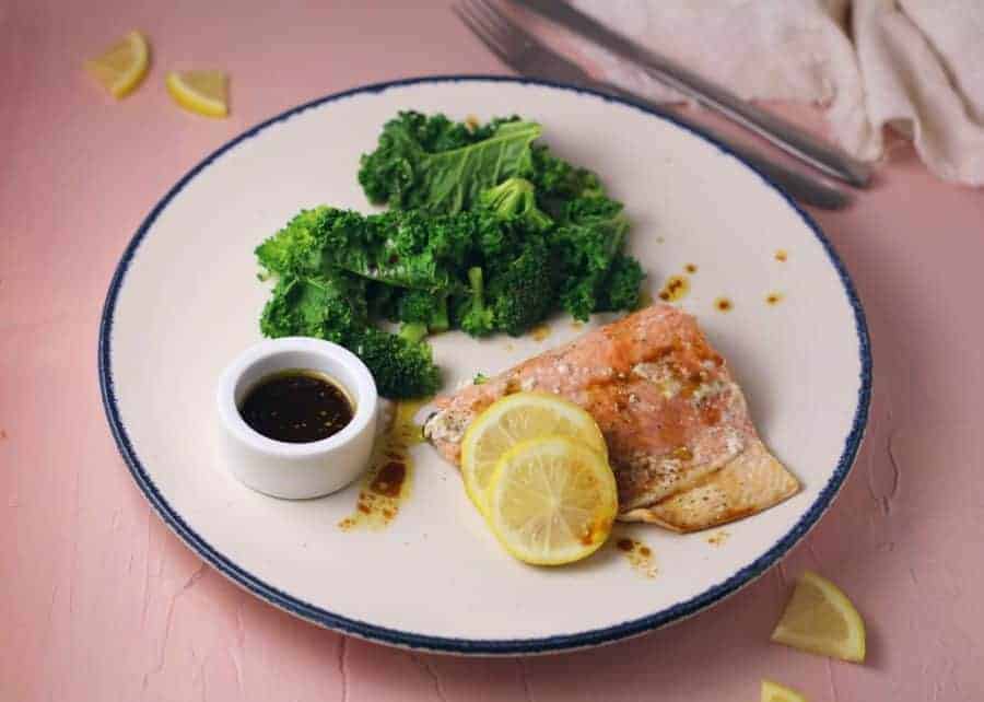 Salmon topped with lemon slices, beside broccoli and brown sauce on a white plate with a blue rim beside cutlery.