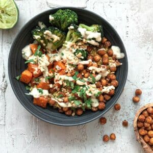 Roasted chickpeas, diced sweet potato and steamed broccoli in grey bowl on white background surrounded by roasted chickpeas and juiced lime.