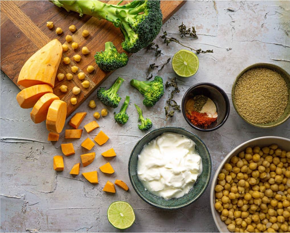 Ingredients in the chickpea Mediterranean quinoa bowl displayed on a wood cutting board.  These include diced potato, roasted chickpeas, lime, broccoli, spices, quinoa and tahini.