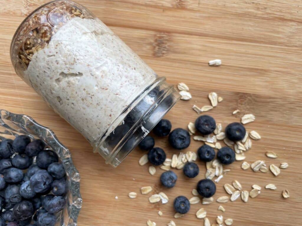 Blueberry cheesecake overnight oats tipped on side with blueberries and oats falling out on a wood board beside a bowl of blueberries.
