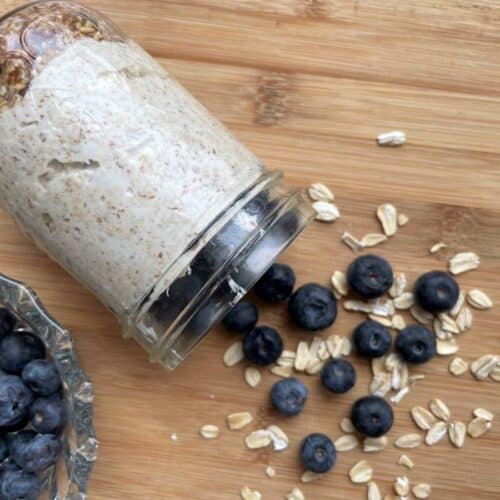 Blueberry cheesecake overnight oats tipped on side with blueberries and oats falling out on a wood board beside a bowl of blueberries.