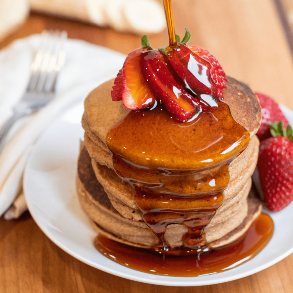 A stack of pancakes on a white plate topped with sliced strawberries  and maple syrup.