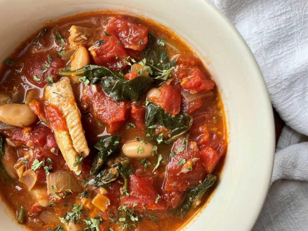 Sardine soup in a white bowl filled with diced tomato, wilted spinach and topped with parsley.