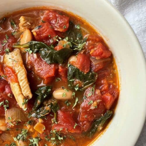 Sardine soup in a white bowl filled with diced tomato, wilted spinach and topped with parsley.